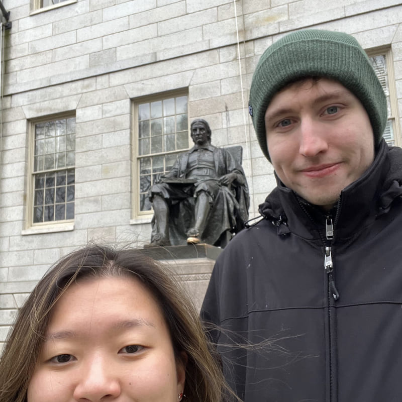 A selfie of a couple in front of a statue of John Harvard